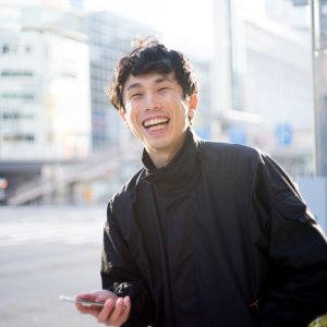 Portrait of Japanese man standing with smile in bright backlight.
He has a smartphone in his hand.